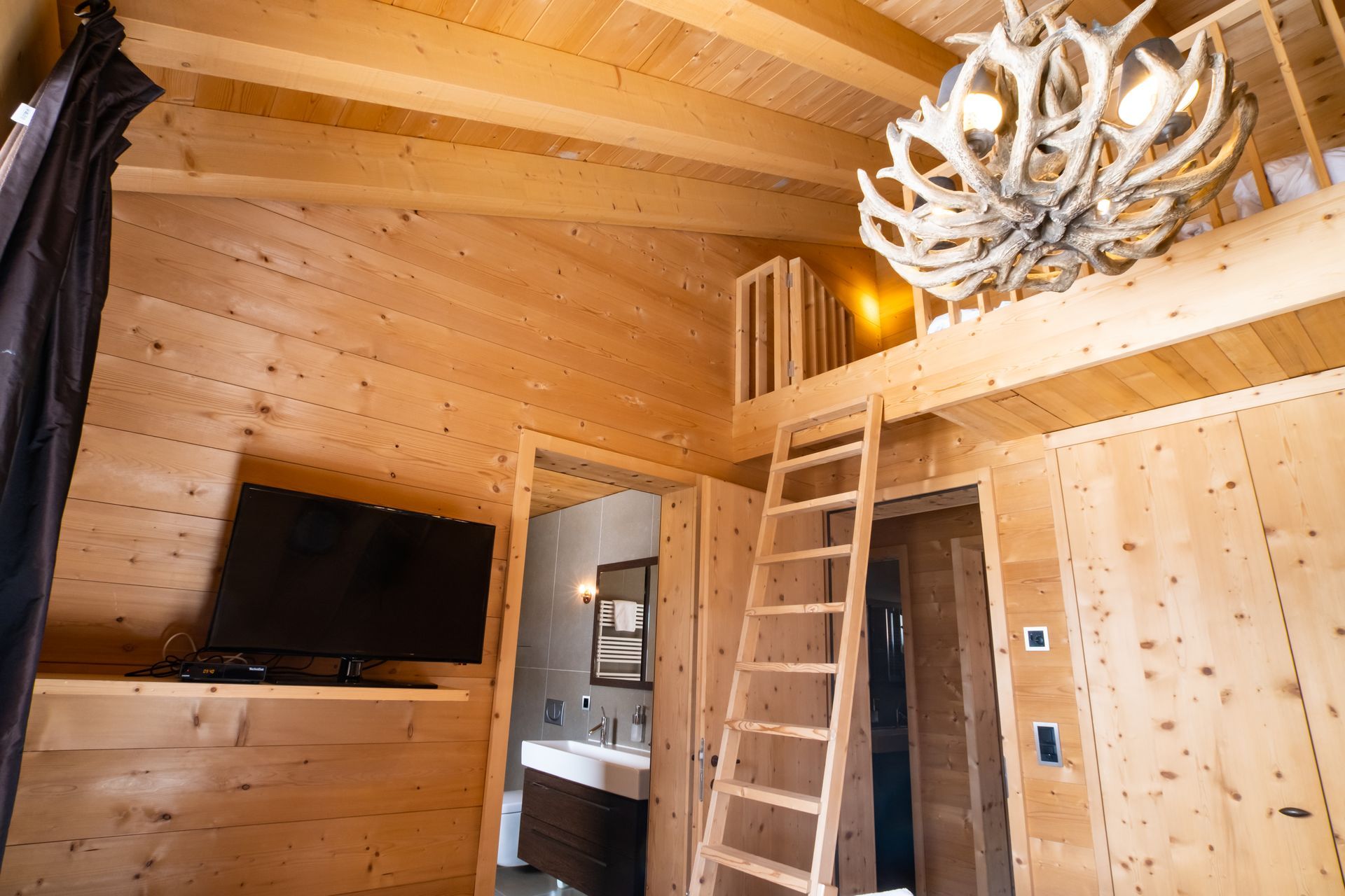 Innenausstattung einer Holzhütte mit Schlafboden, Fernseher, Leiter und Kronleuchter aus Geweih.