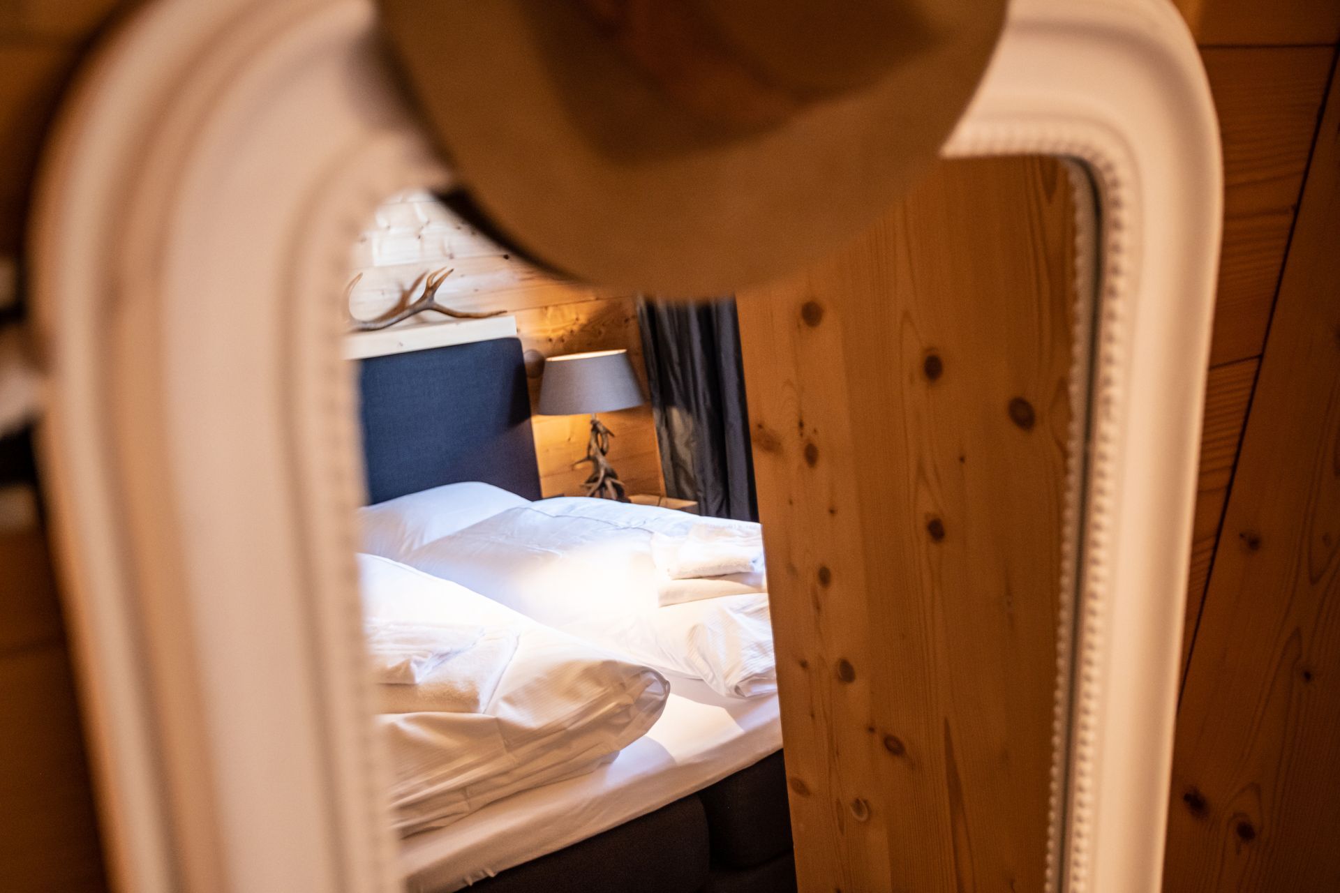 Ein Spiegel reflektiert ein Schlafzimmer mit Bett, Lampe und Hut. Holzwände und eine neutrale Farbgestaltung.