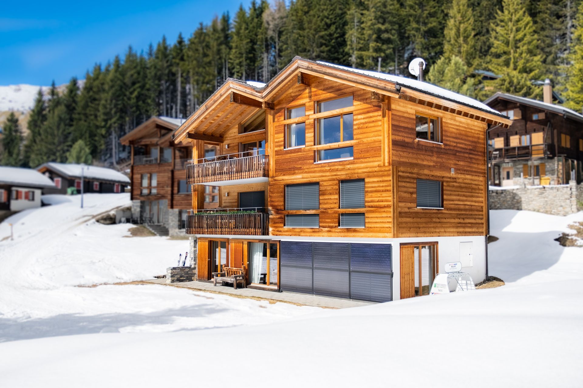 Holzhäuser im Chalet-Stil in einer verschneiten Landschaft vor einem Waldhintergrund. Sonniger Tag.