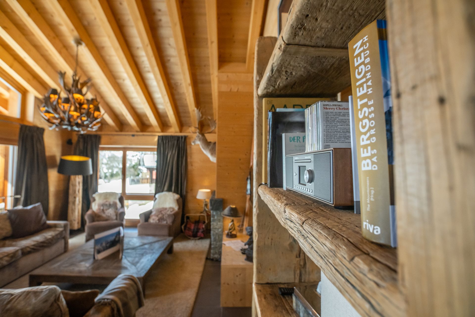Gemütliches Wohnzimmer mit Holzbalken und Bücherregal. Sonnenlicht fällt durch das Fenster und erhellt die Sitzecke und den Kronleuchter.