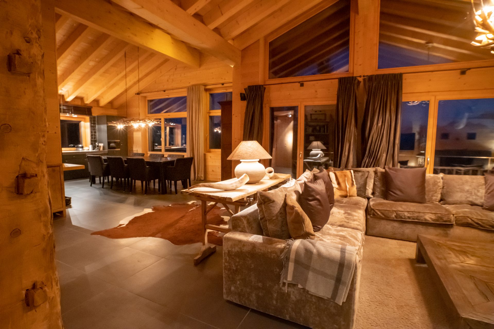 Gemütliches Wohnzimmer mit Holzbalken, großen Fenstern und einem braunen Sofa. Im Hintergrund der Esstisch.