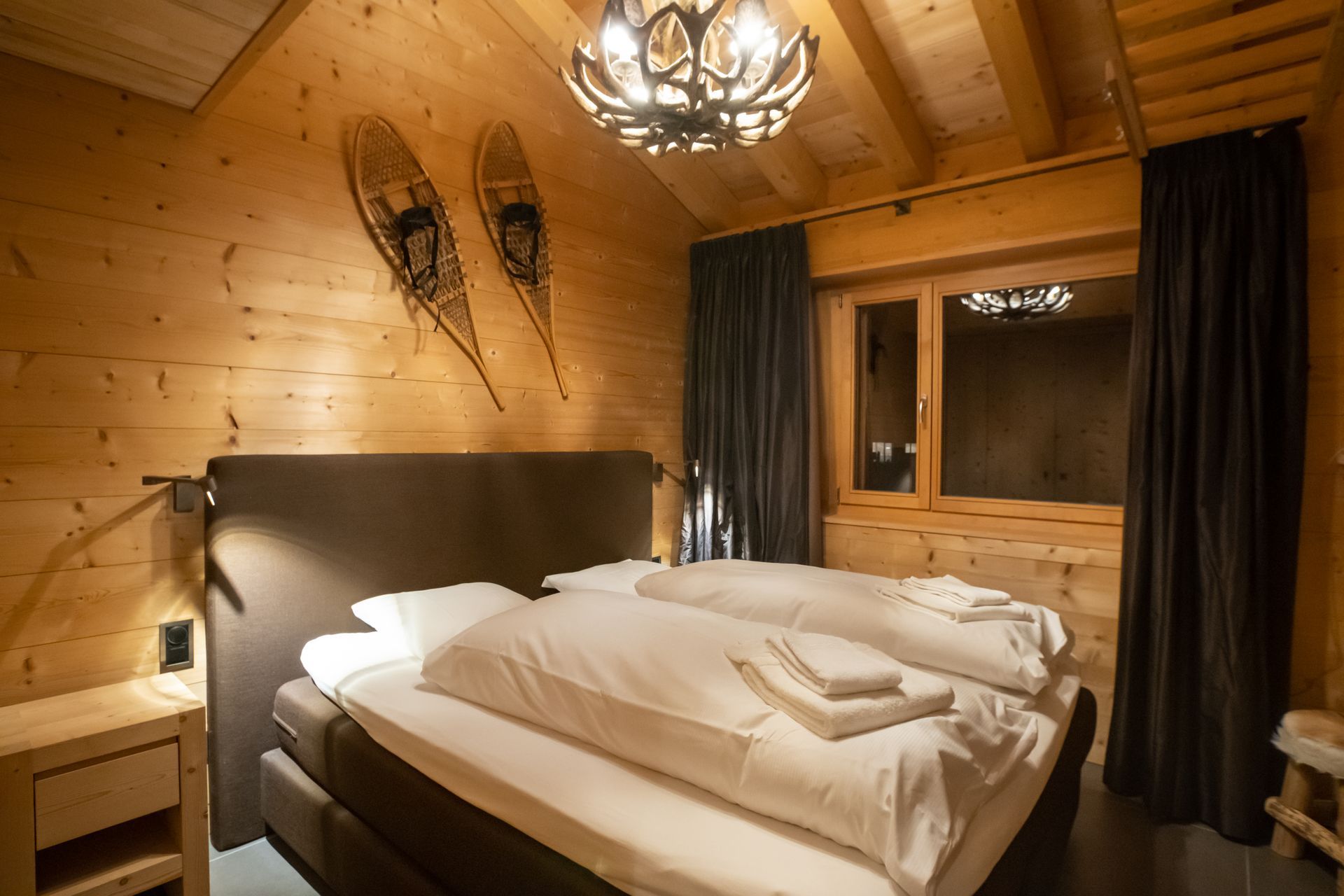 Gemütliches Schlafzimmer mit Holzwänden, Bett mit weißer Bettwäsche, Schneeschuhdekor, Kronleuchter und einem Fenster mit dunklen Vorhängen.