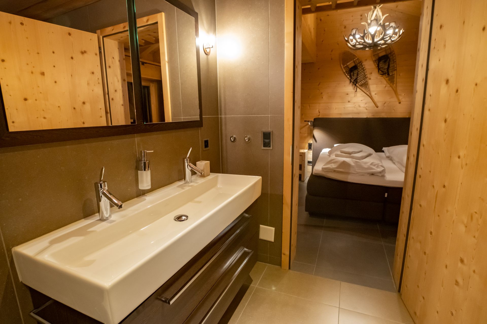 Badezimmer mit einem langen weißen Waschbecken, Spiegel und Tür zu einem Schlafzimmer mit Bett.