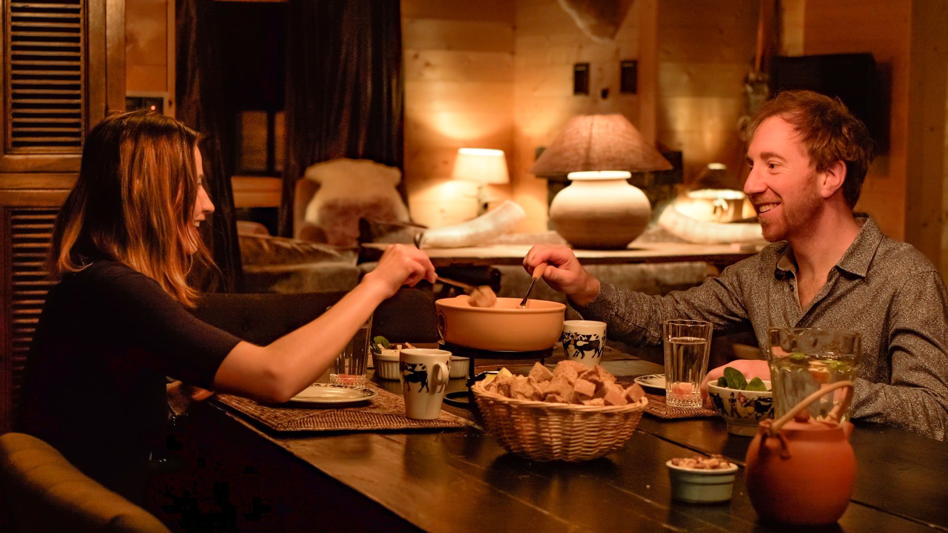Ein Paar bei einem Abendessen im Kerzenschein, beide greifen nach Essen aus einer Schüssel auf einem dunklen Holztisch.
