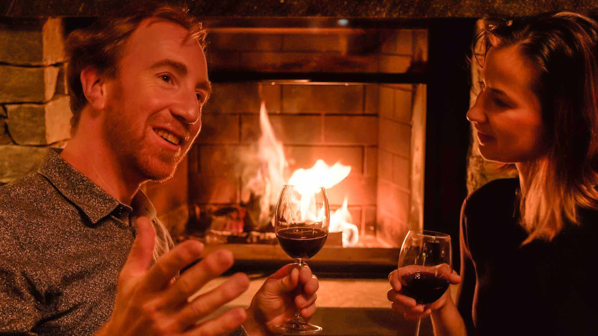 Ein Paar stößt vor einem Kamin mit Wein an und genießt einen gemütlichen Abend.