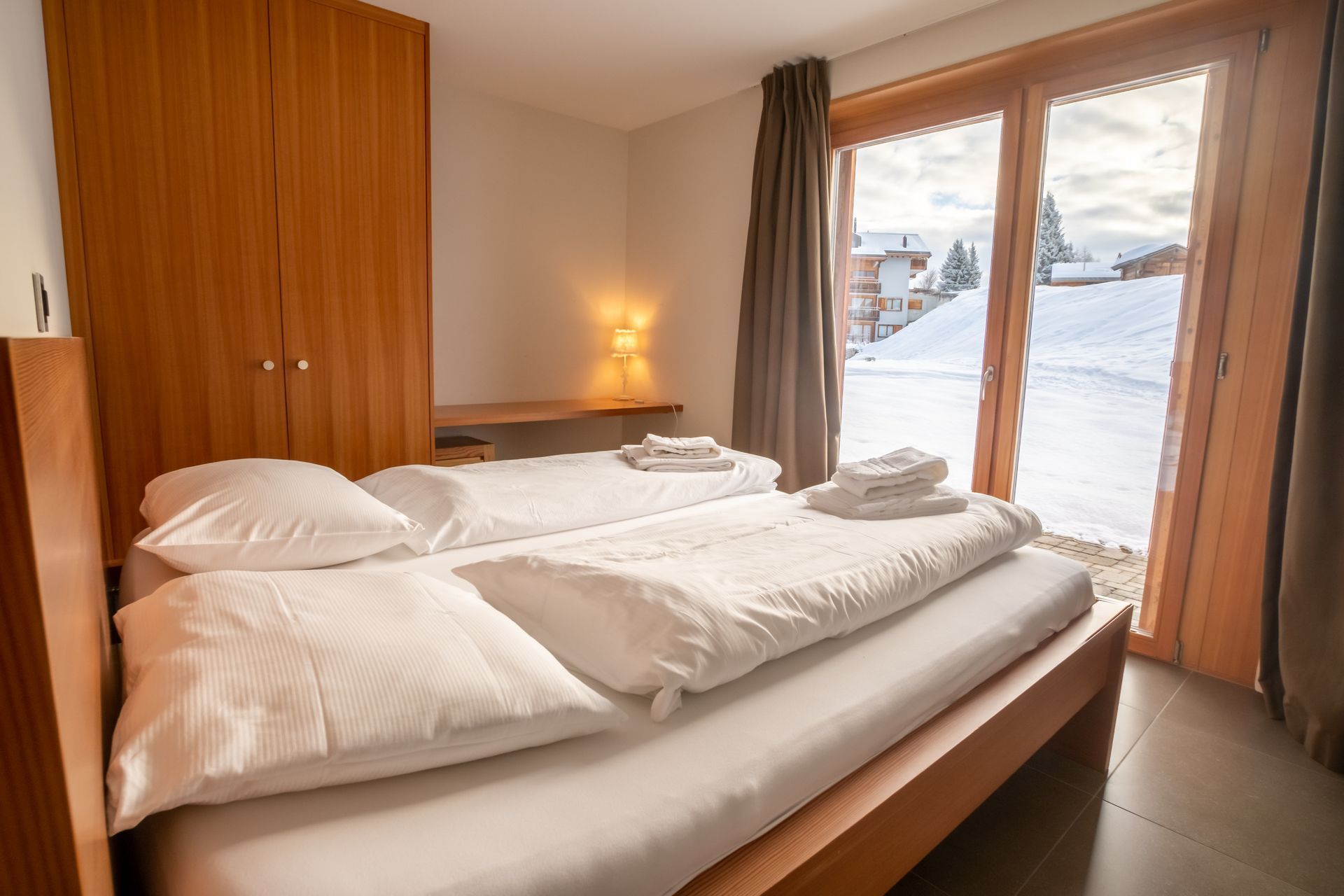 Schlafzimmer mit Holzbett, weißer Bettwäsche und großen Fenstern mit Blick auf eine verschneite Landschaft.