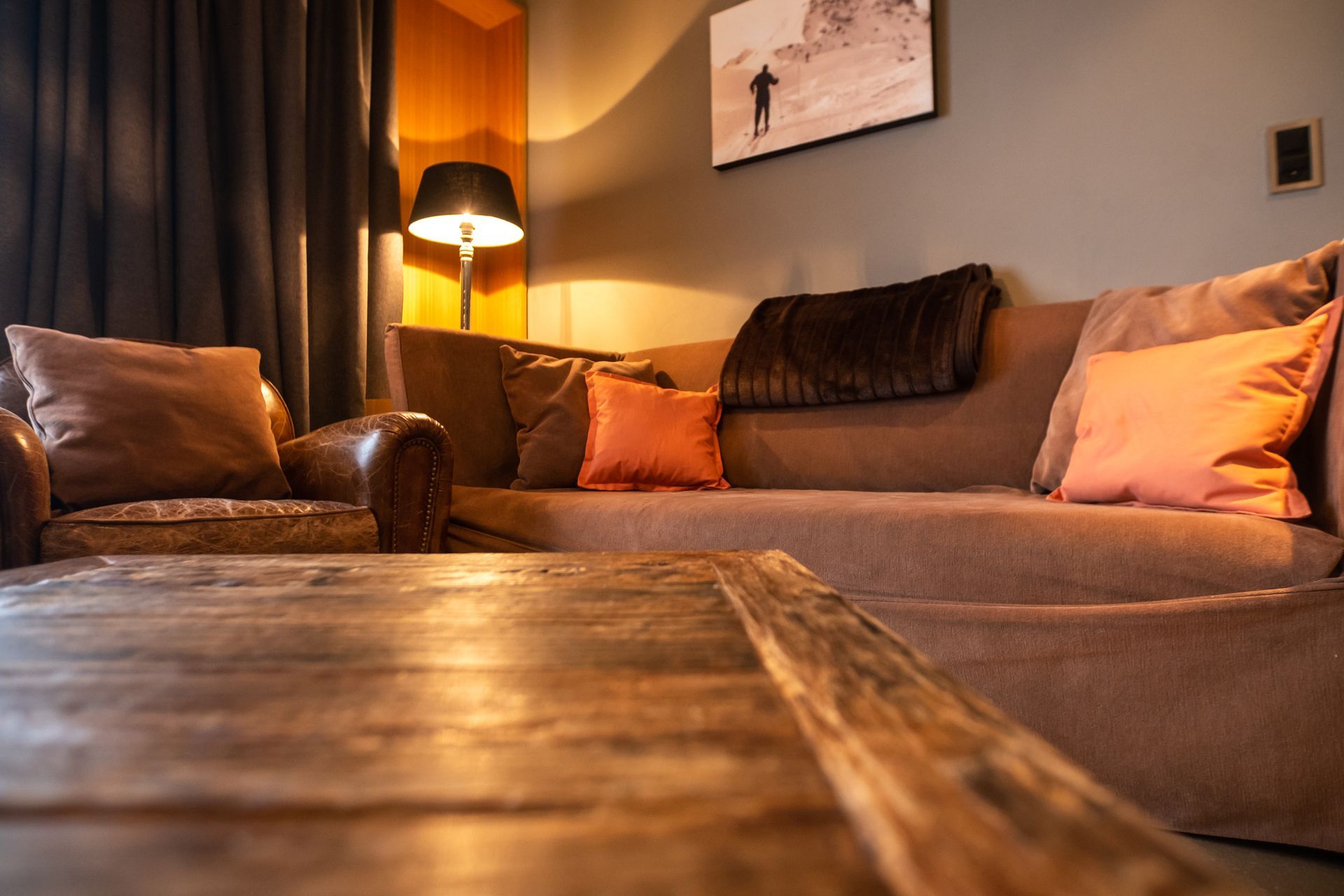 Gemütliches Wohnzimmer mit braunem Sofa, Ledersessel, orangefarbenen Kissen und Couchtisch aus Holz.