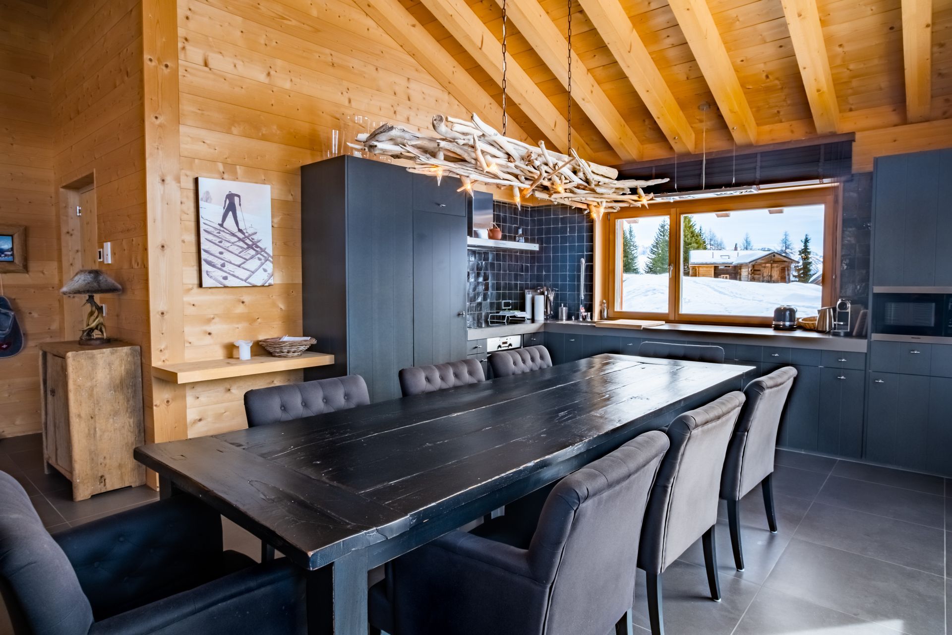 Esszimmer mit langem schwarzem Tisch, grauen Stühlen, Küche mit Schnee draußen vor dem Fenster.