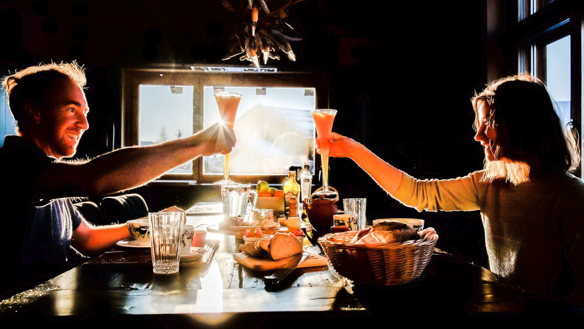 Ein Paar stößt mit Getränken an einem Esstisch in der Nähe eines sonnenbeschienenen Fensters an.