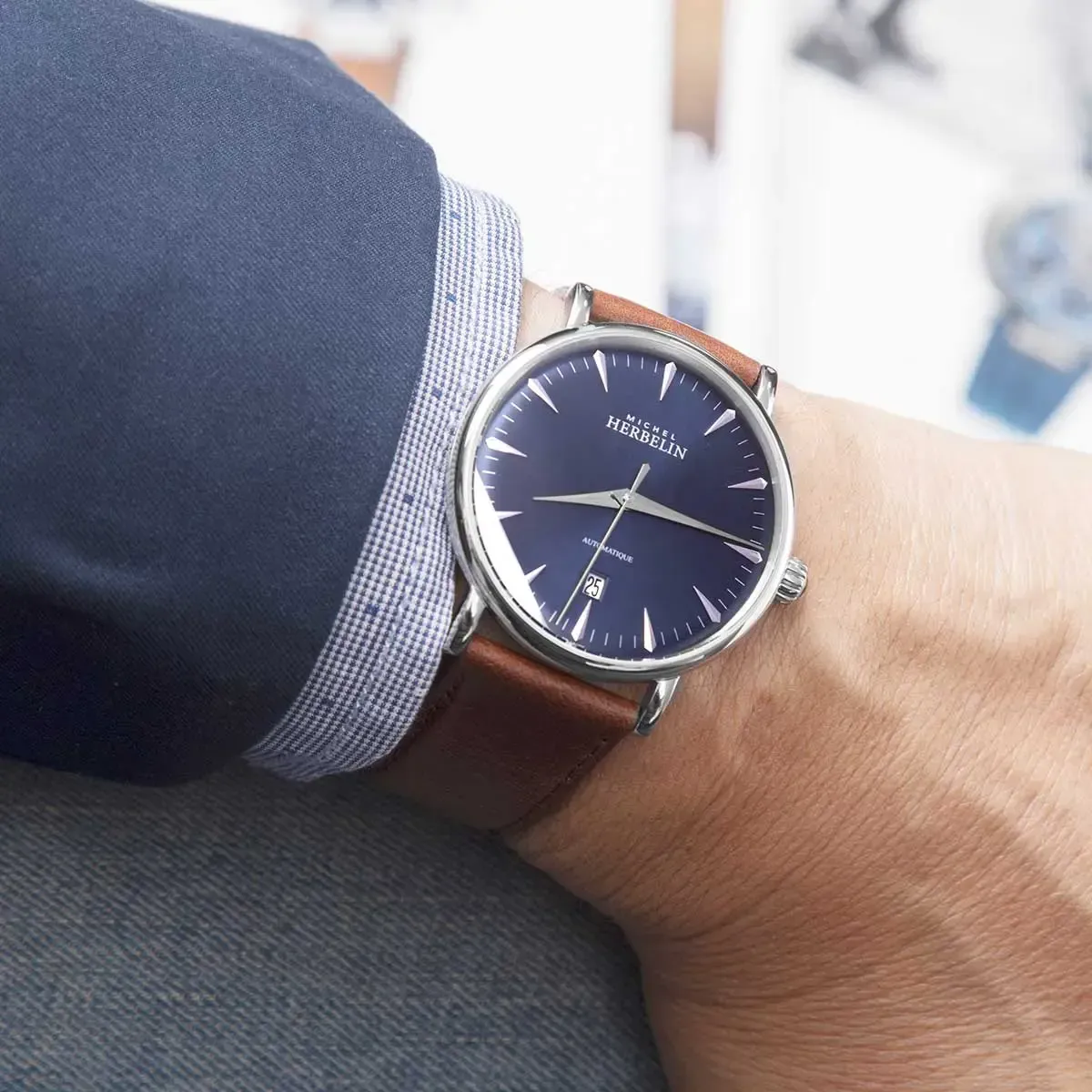 Homme portant une montre-bracelet en argent avec un cadran bleu et un bracelet en cuir marron, visible sur une manche de blazer bleu.