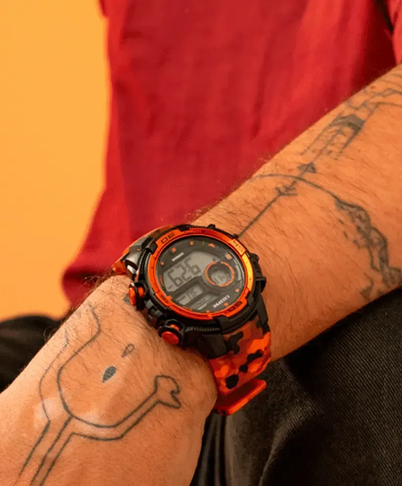 Bras d'homme avec montre numérique orange et noire, tatouages ​​visibles, chemise rouge, fond orange.