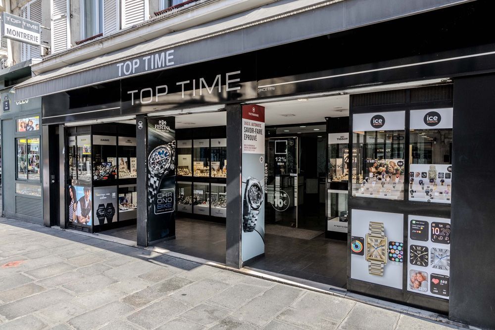 Une devanture de magasin avec un auvent noir sur lequel est écrit TOP TIME présente des montres ; une femme est sur une bannière.