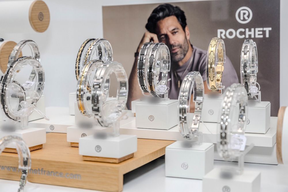 Des horloges en cristal sont exposées sur des supports blancs dans une salle d'exposition, avec un homme en arrière-plan près du logo ROCHET.