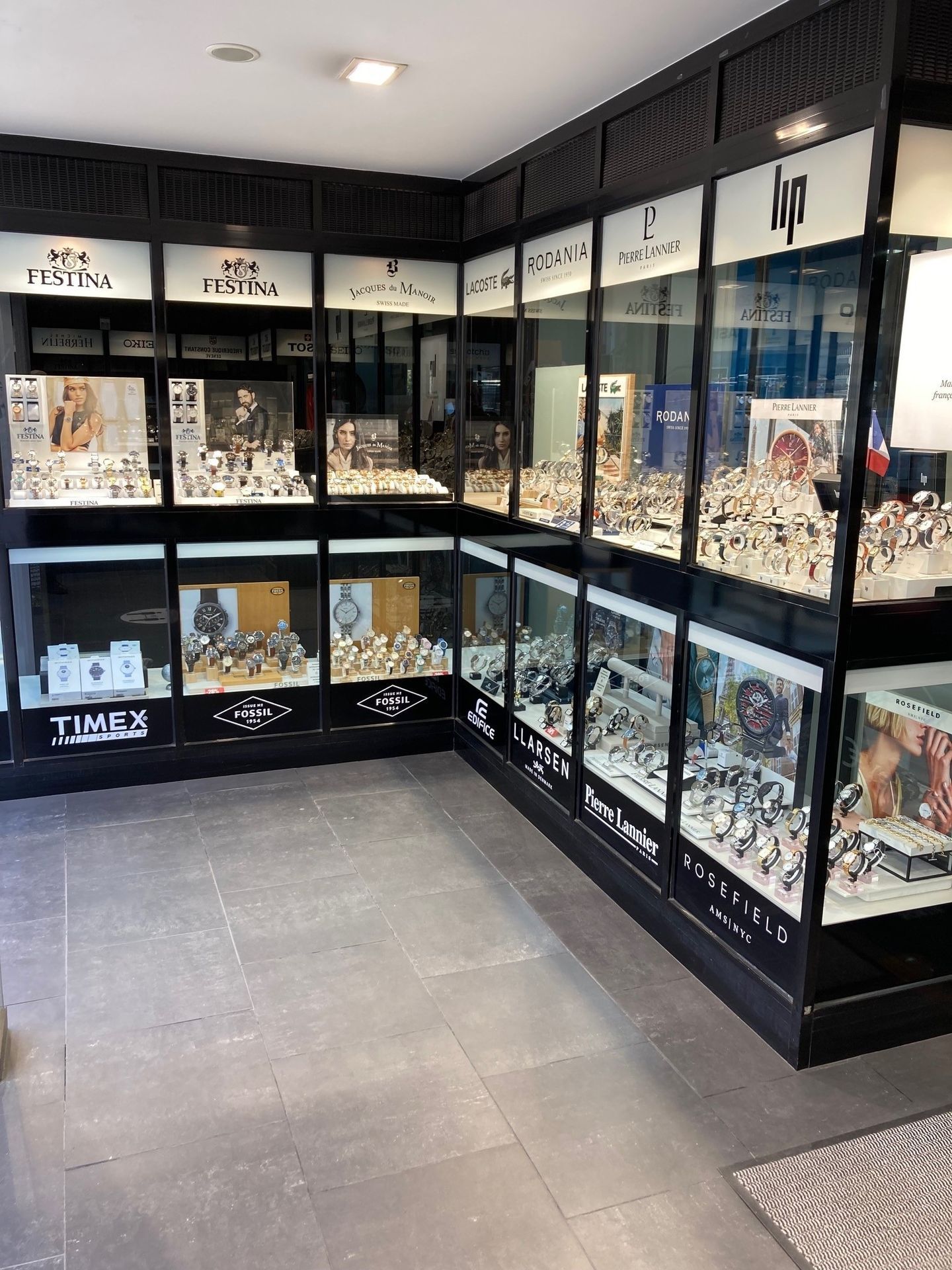Intérieur d'un magasin de montres avec vitrines remplies de montres ; sol en carrelage gris.