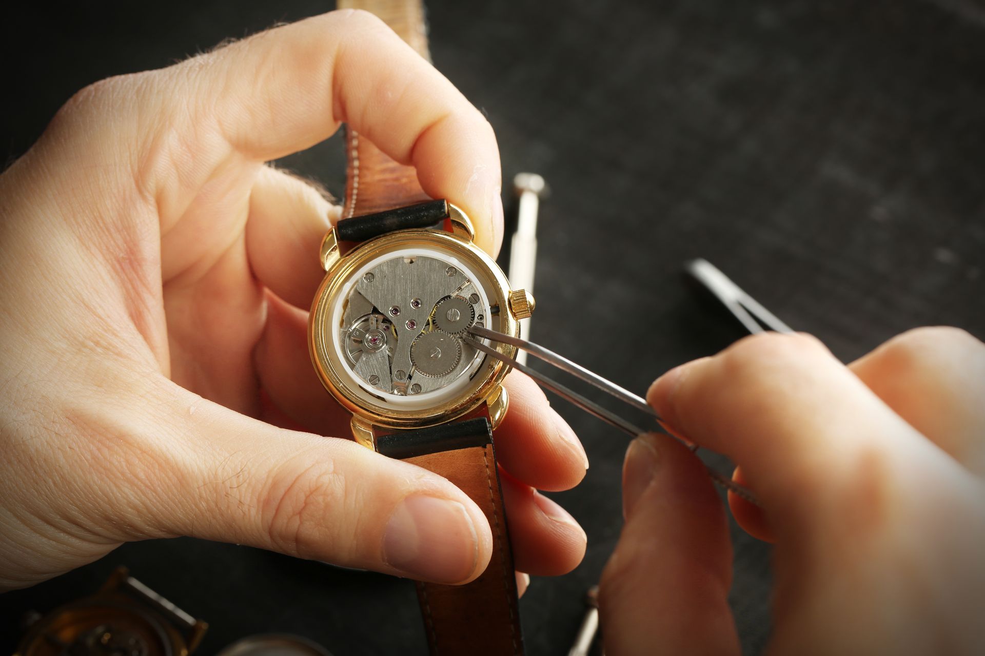 Une personne utilise une pince à épiler pour réparer le fonctionnement interne d'une montre-bracelet en or, sur une surface noire.