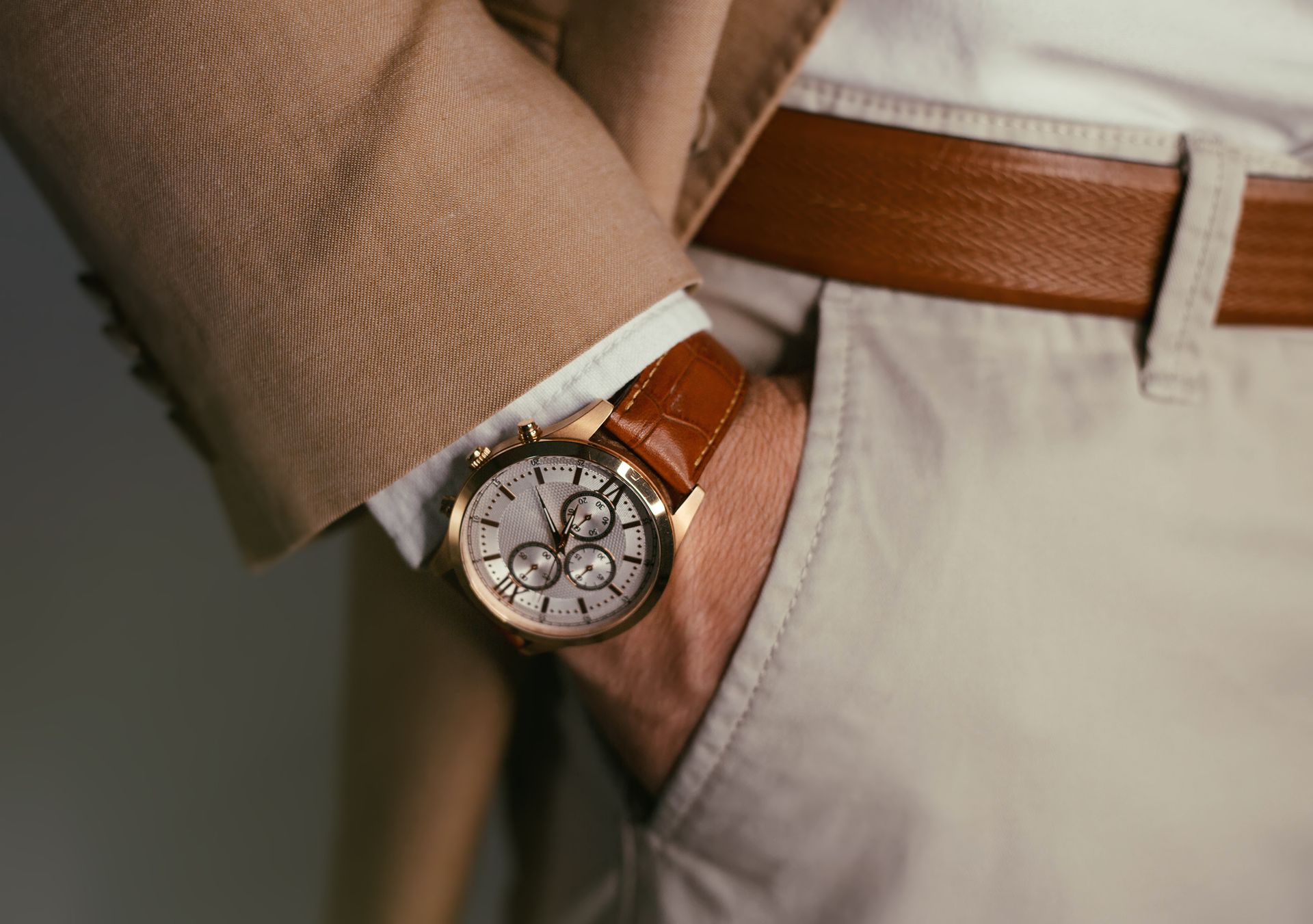 Bras d'homme avec montre en or, main dans la poche, portant une veste beige, une ceinture beige et un pantalon clair.