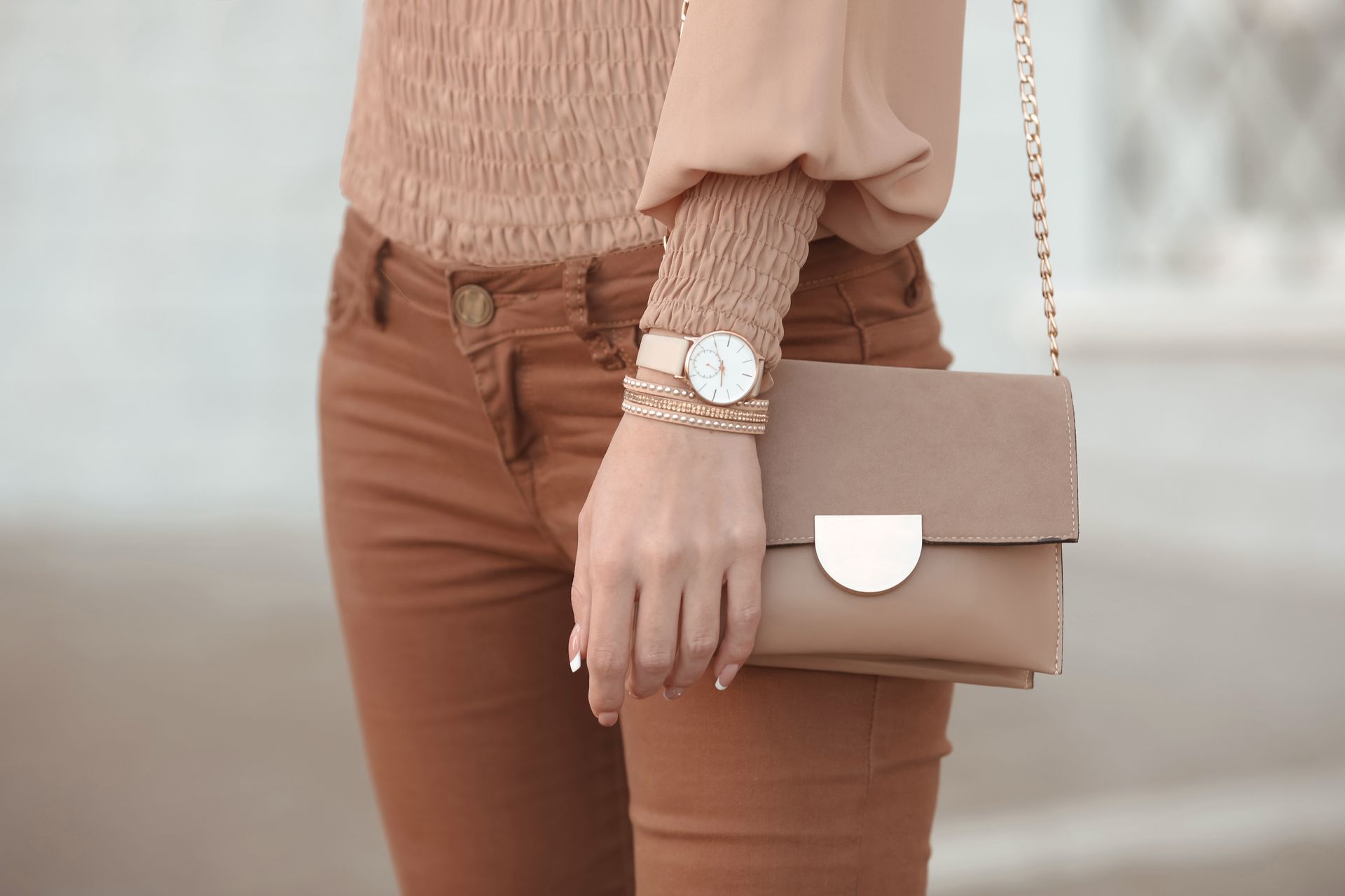 Femme en tenue beige, tenant un sac à main, portant une montre, debout à l'extérieur.