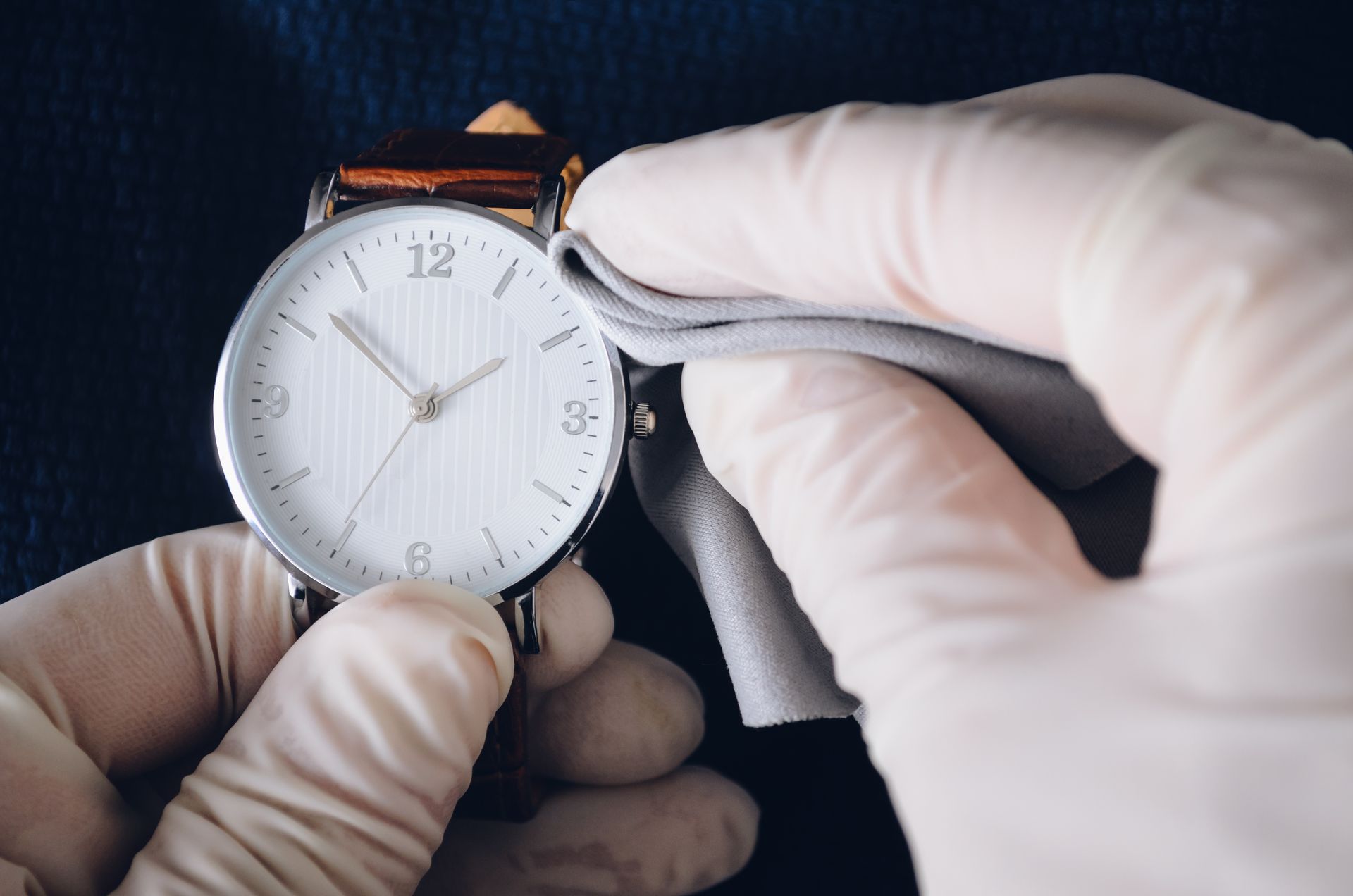 Mains gantées nettoyant une montre-bracelet en argent avec un chiffon gris. La montre a un cadran blanc et un bracelet en cuir marron.