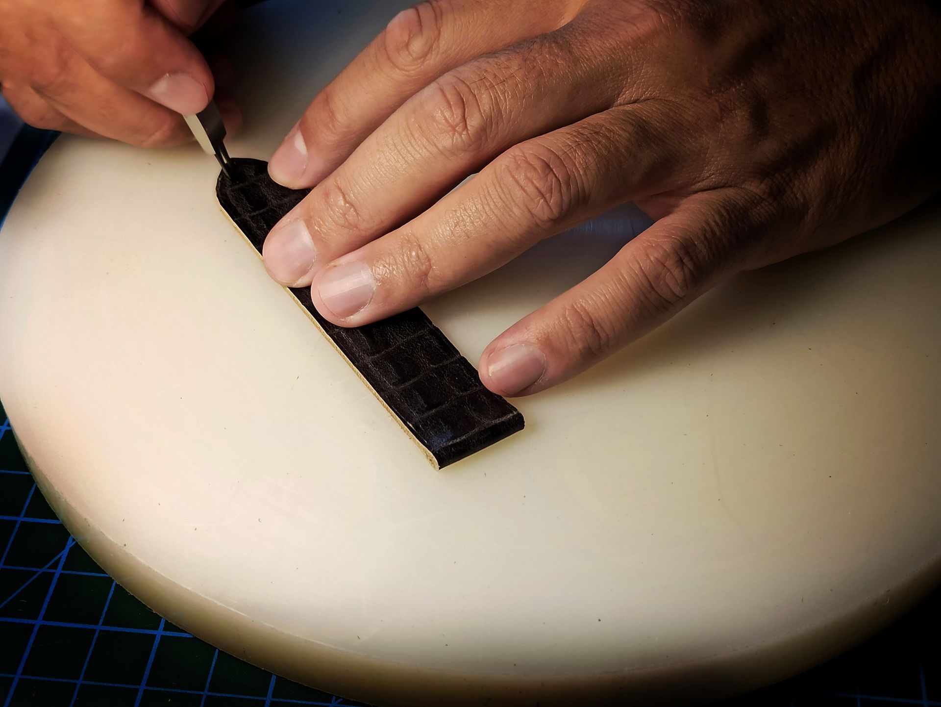 Mains utilisant un outil sur une bande de cuir noir, probablement pour l'artisanat, sur une surface blanche.