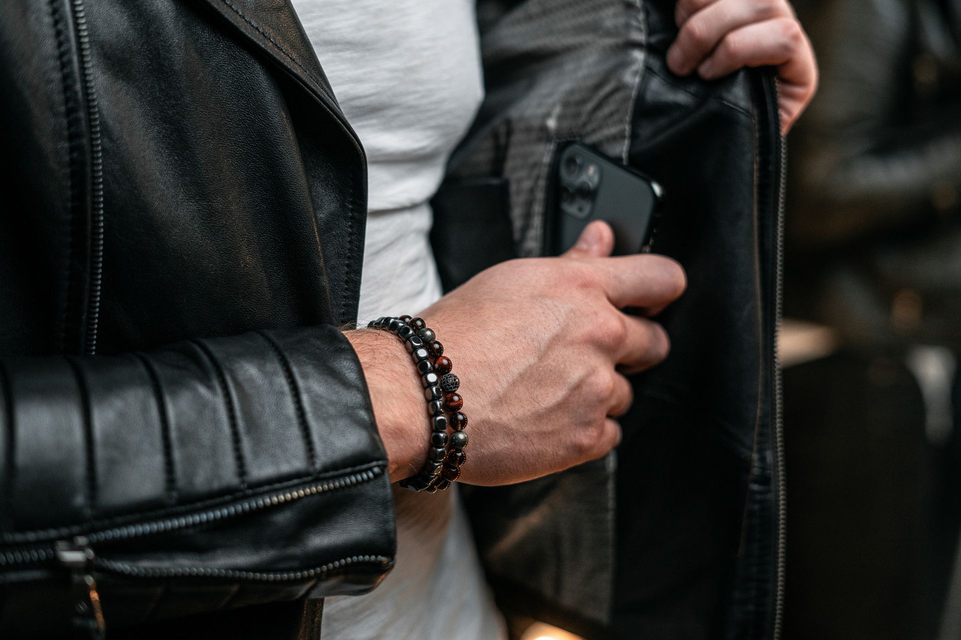 Homme en veste de cuir noir, rangeant son téléphone dans sa poche intérieure. Bracelet noir au poignet.