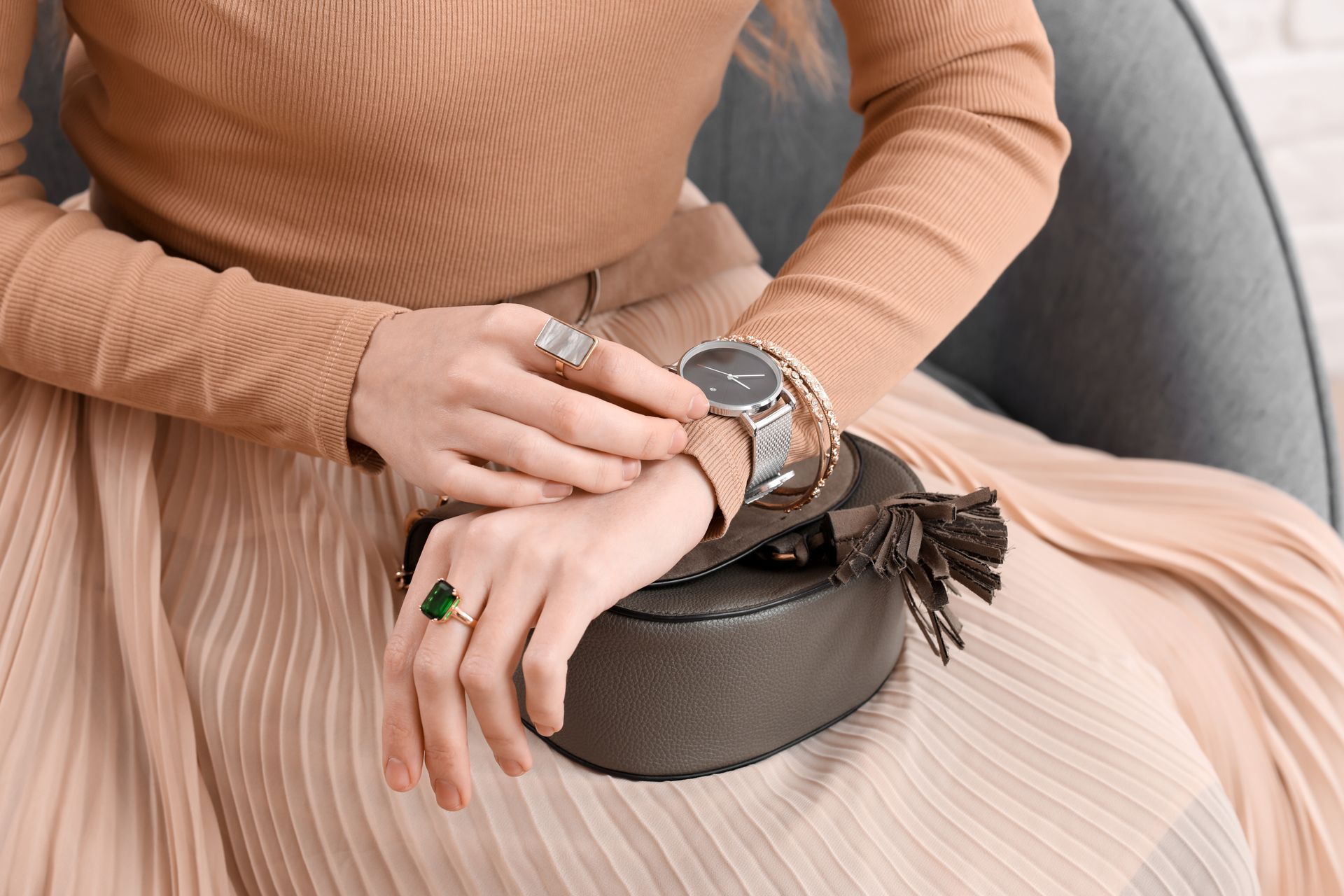Femme portant des bijoux, les mains posées sur un sac à main, vêtue d'une jupe plissée, tons neutres.
