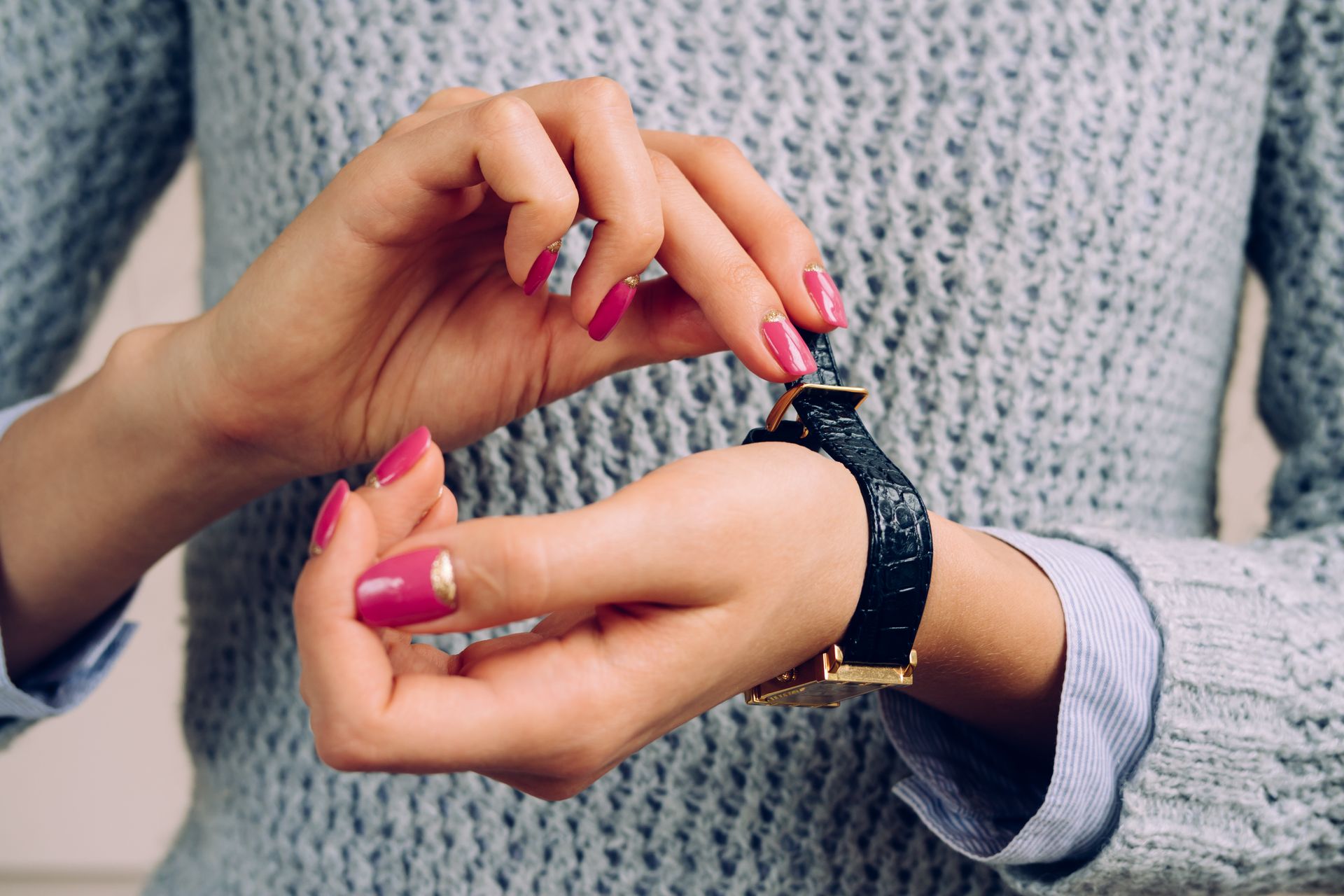 Mains d'une femme ajustant une montre noire avec des détails dorés, portant un pull gris, ongles peints en rose avec des motifs dorés.