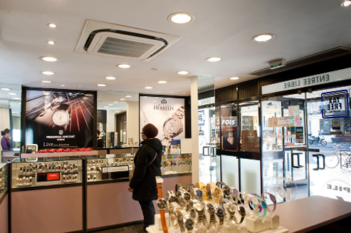 Une femme dans un magasin de montres regarde les présentoirs ; des montres, des miroirs et des affiches promotionnelles sont visibles.