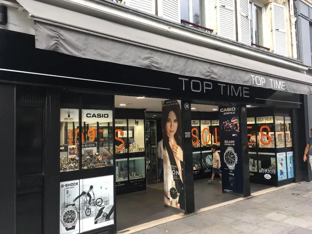 Une devanture de magasin avec un auvent noir sur lequel est écrit TOP TIME présente des montres ; une femme est sur une bannière.