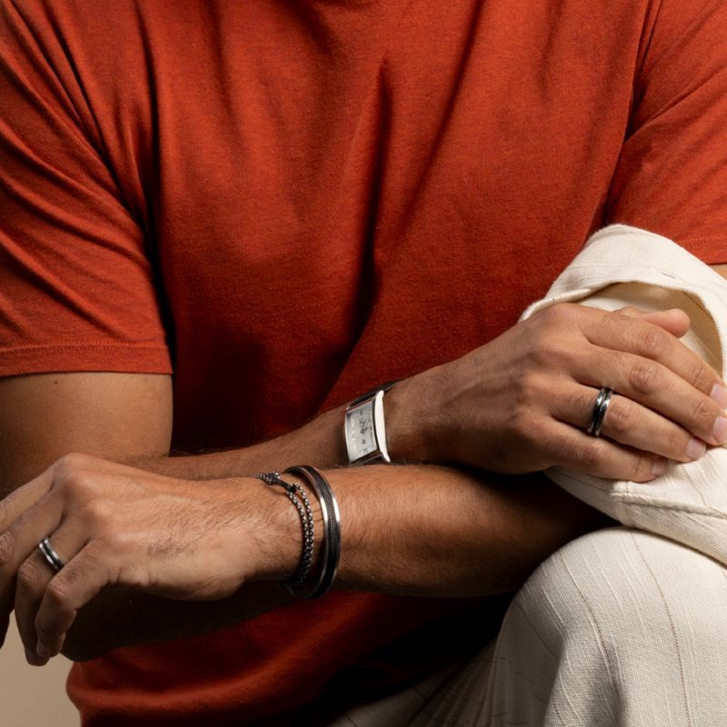 Homme portant des bagues, une montre et des bracelets, les mains posées sur un tissu crème, avec un T-shirt couleur rouille.