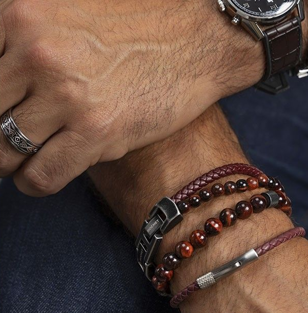 Bras d'homme portant une montre, une bague et trois bracelets ; tons rouge/marron.