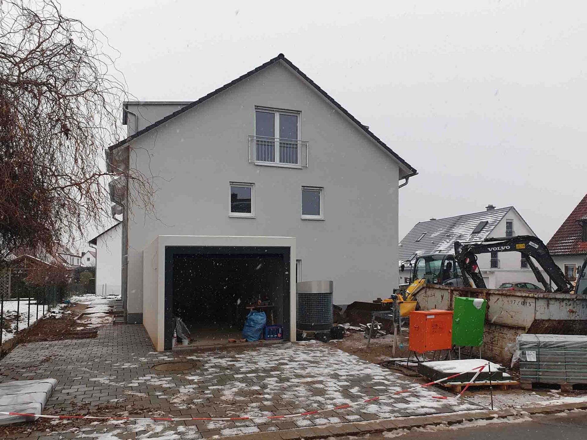 Neu gebautes Haus mit offener Garage und Baumaschinen im Hof, verschneiter Boden.
