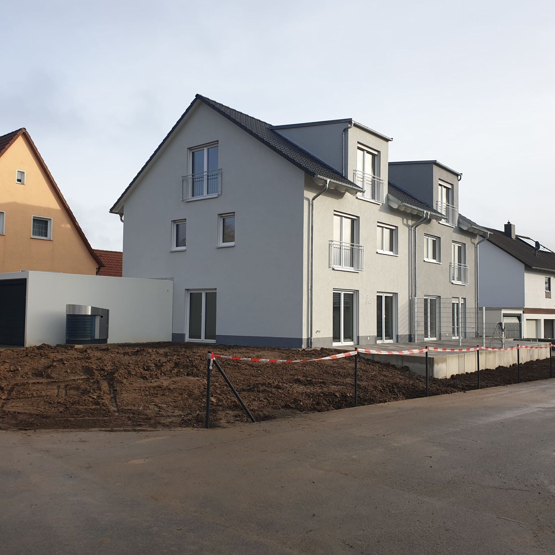 Mehrstöckiges graues Haus mit Dachgauben, neu gebaut, unbefestigtes Grundstück im Vordergrund.