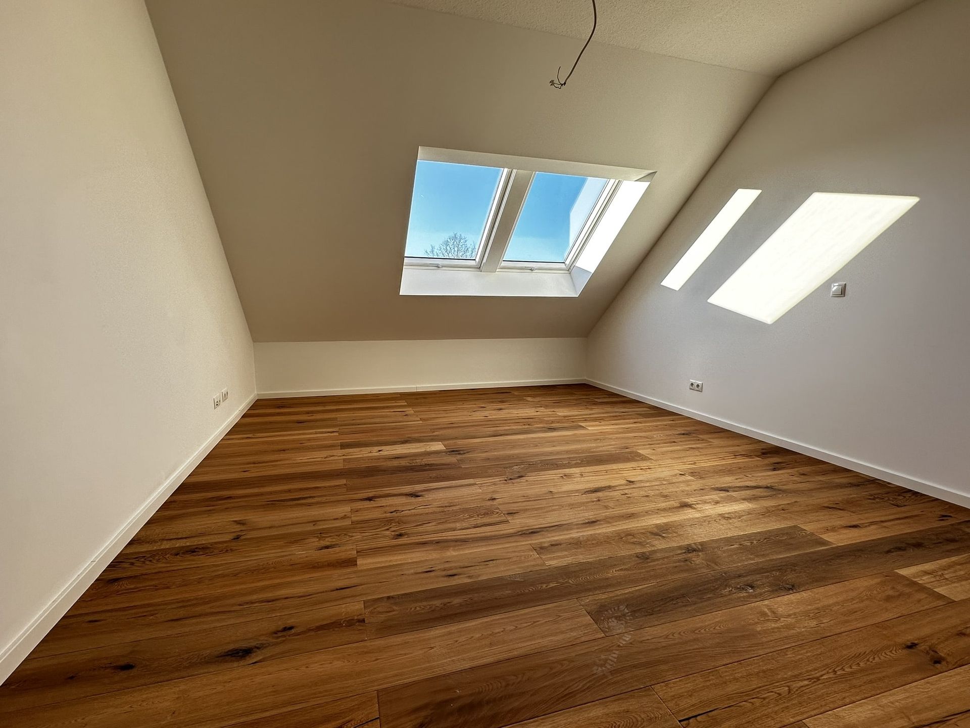 Leerer Raum mit schrägen weißen Wänden, Holzboden und Oberlicht. Sonnenlicht strömt herein.