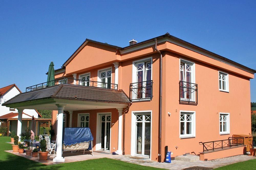 Zweistöckiges orangefarbenes Haus mit weiß gerahmten Fenstern, einer überdachten Terrasse und einem Rasen unter blauem Himmel.