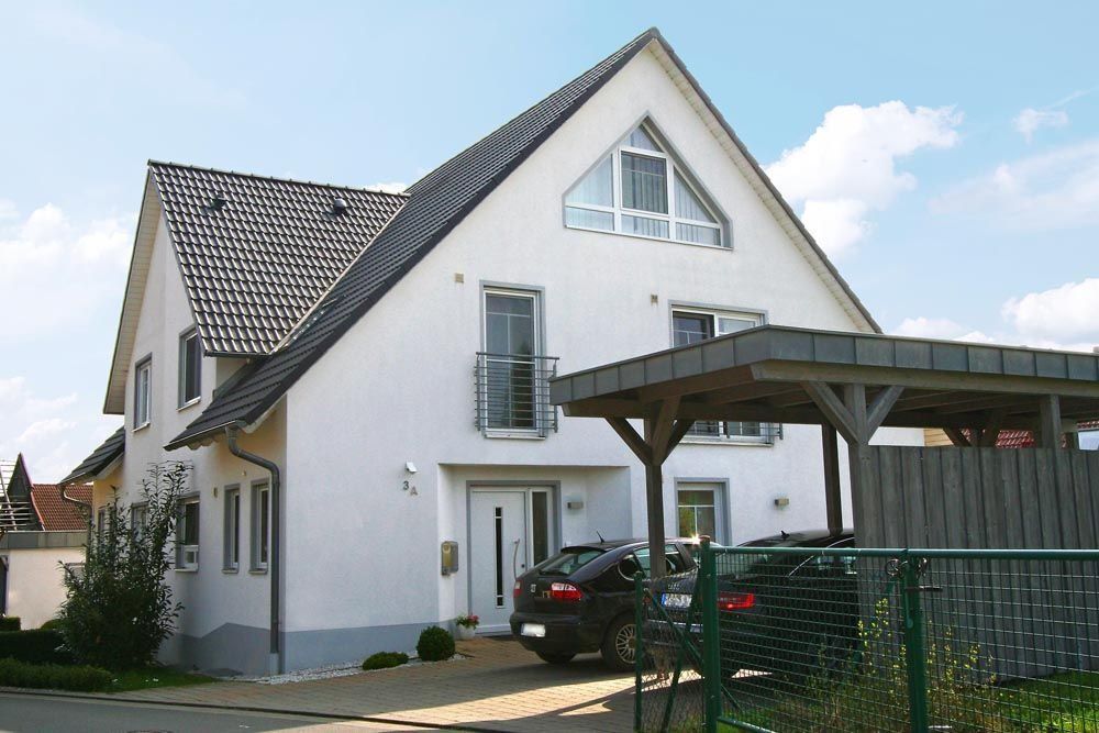 Weißes zweistöckiges Haus mit schwarzem Ziegeldach und Carport, in der Einfahrt parkt ein Auto.