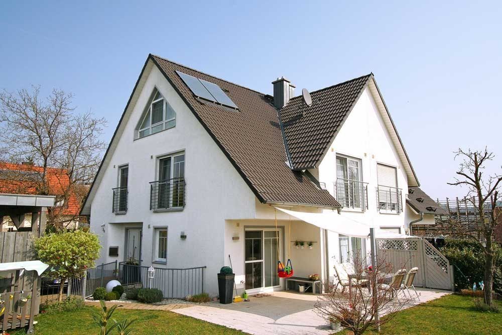 Weißes Haus mit dunklem Dach, Balkonen und Sonnenkollektoren an einem sonnigen Tag.