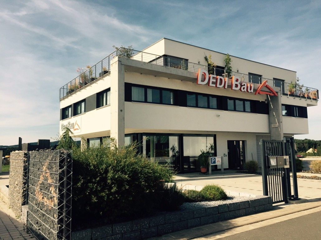 Modernes, weißes Gebäude mit „DEDI BAU“-Schild. Grauer Steinzaun davor, blauer Himmel.