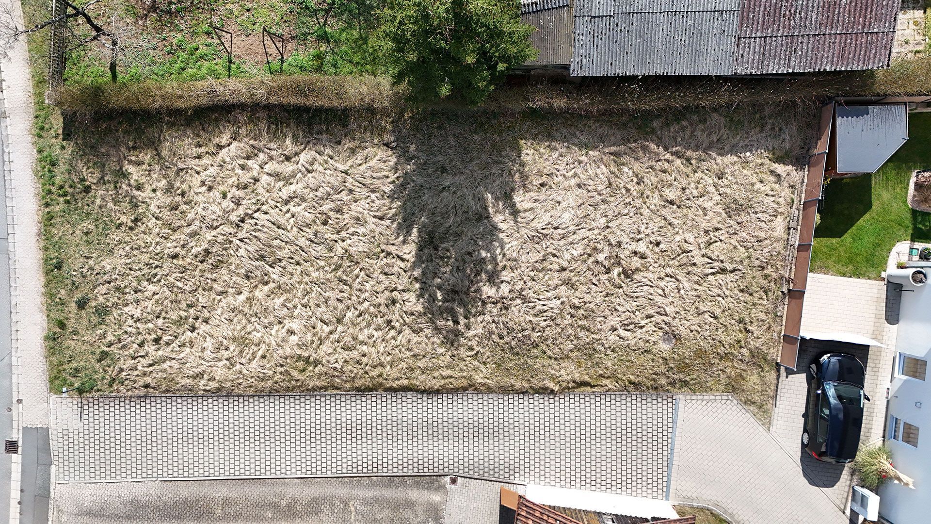 Draufsicht auf einen rechteckigen Hof mit einer Hecke, einem kleinen Baum und einem Gebäude, neben dem ein Auto geparkt ist.