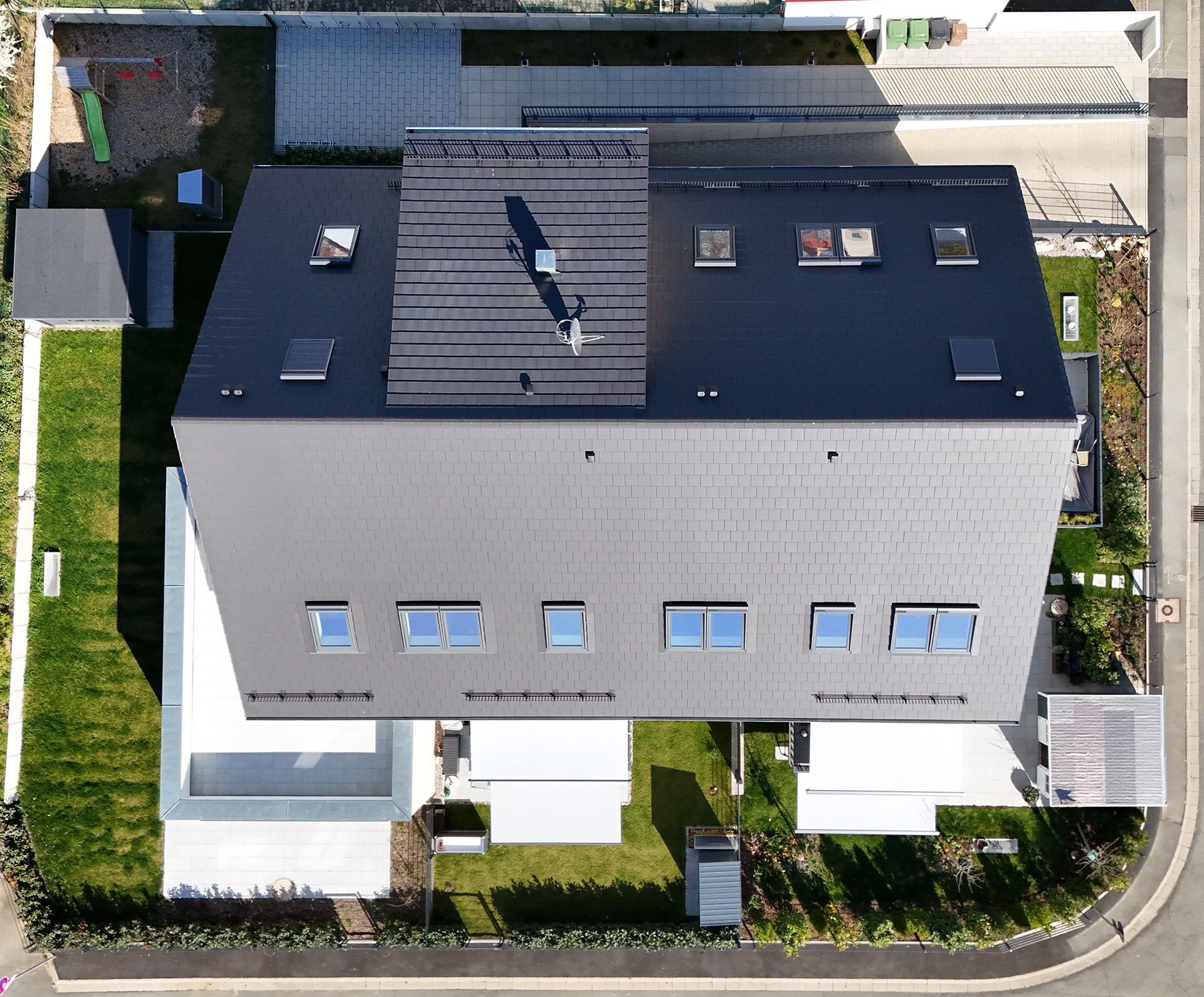 Draufsicht auf ein modernes Haus mit dunklem Dach und umgebendem grünem Rasen und Garten.