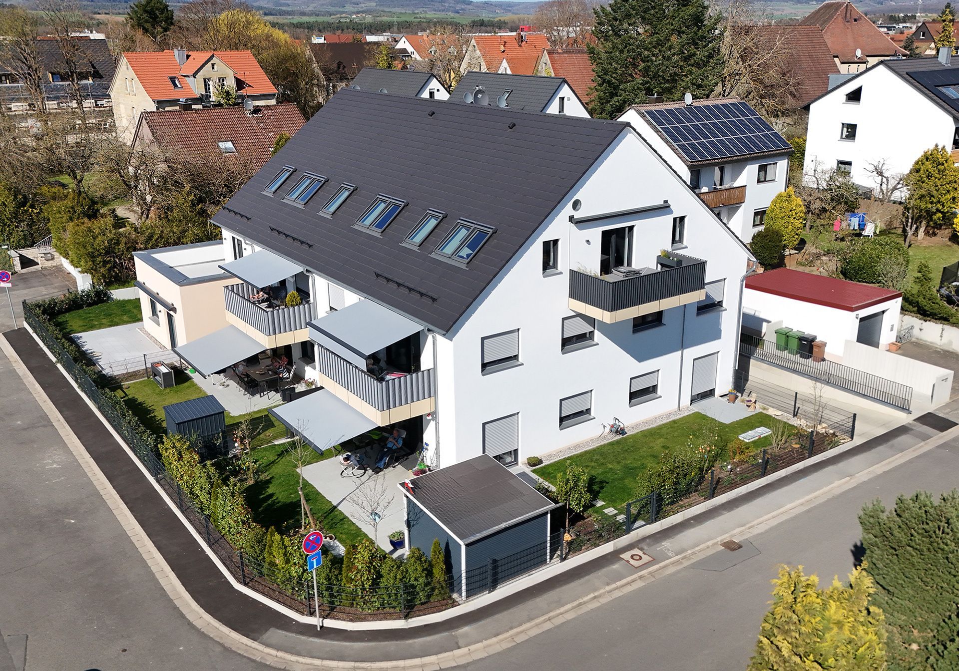 Modernes dreistöckiges Wohnhaus mit dunkelgrauem Dach und Balkonen, umgeben von grünen Rasenflächen und einer Straße.