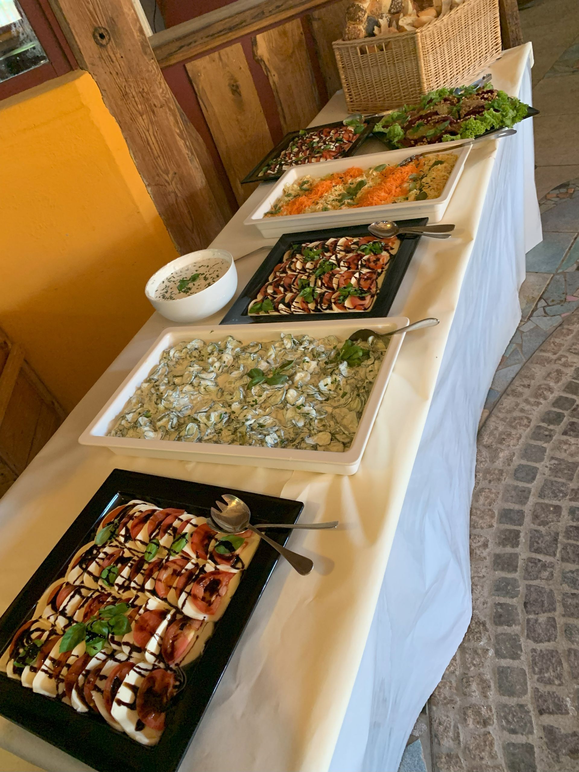 Buffettisch mit verschiedenen Salaten: Caprese, Pasta, Couscous und Gemüse. Holzhintergrund, weiße Tischdecke.