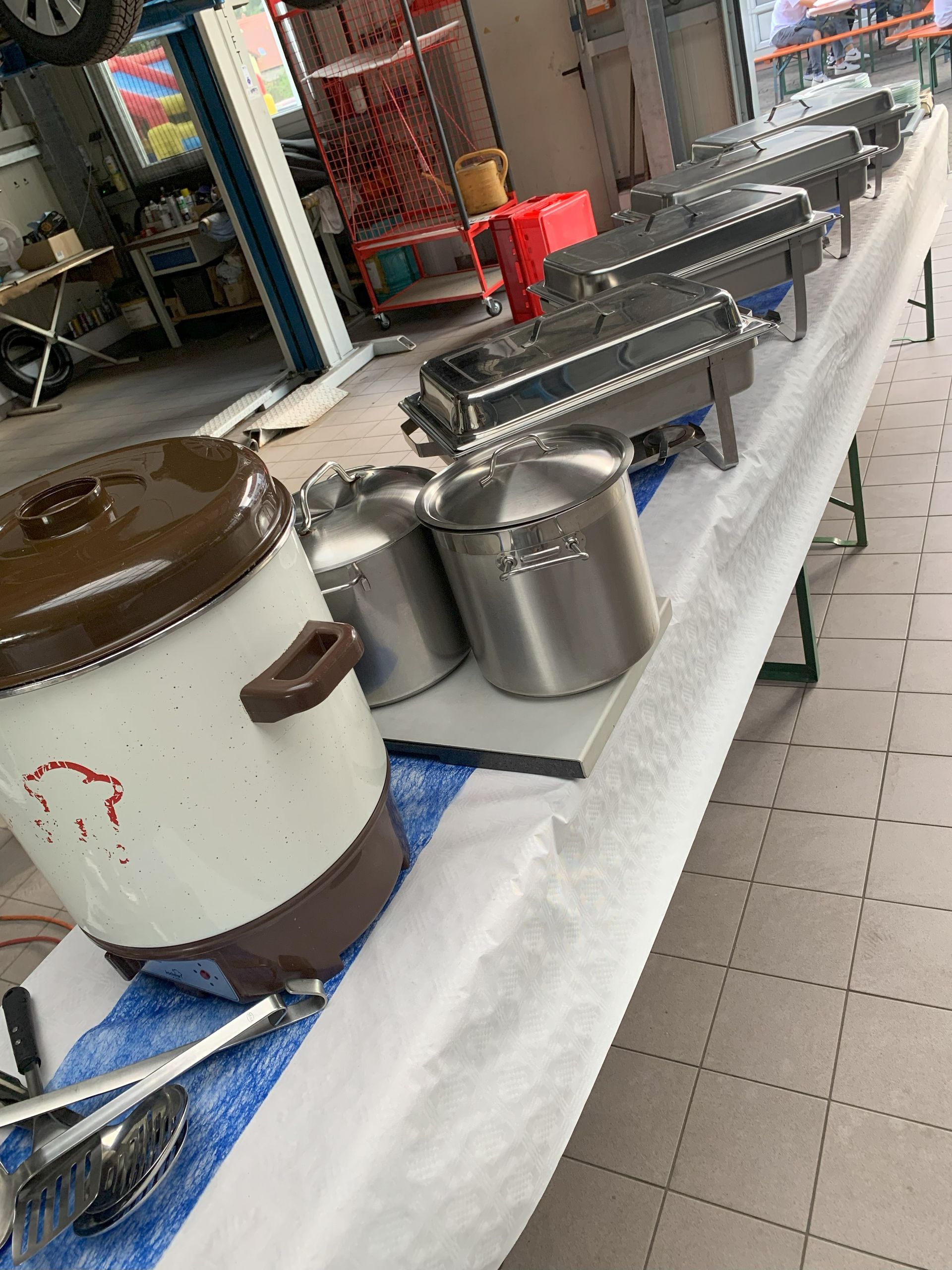 Buffetaufbau mit abgedeckten Lebensmittelbehältern auf einem langen Tisch, wahrscheinlich im Innenbereich, mit Kochausrüstung.