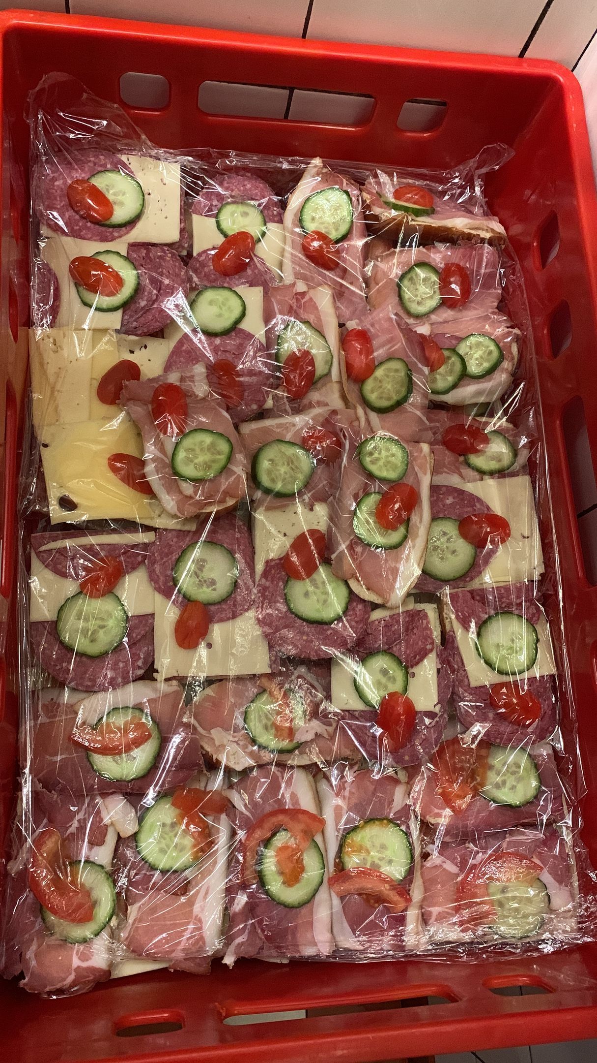 Sandwiches in durchsichtiger Plastikfolie, gestapelt in einer roten Kiste, belegt mit Gurken, Tomaten und verschiedenen Fleisch- und Käsesorten.