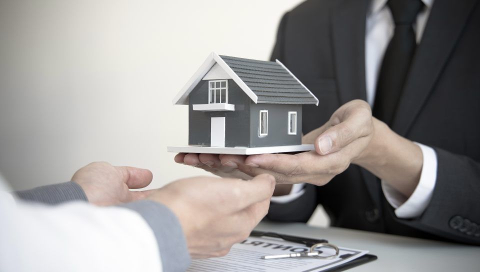 Modellhaus wird von zwei Personen über Tisch zueinander gereicht