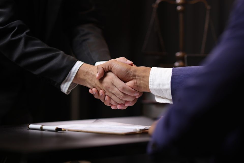 Zwei Personen reichen sich die Hand über Schreibtisch, Dokumentenklemmbrett