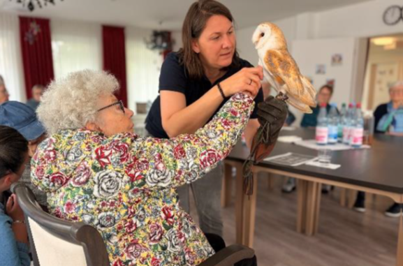 Woman holding an owl while seated woman reaches out in a bright indoor room