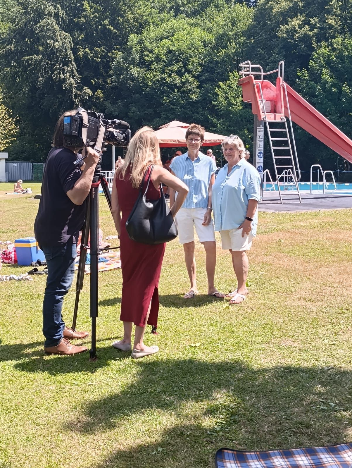 Menschen posieren für ein Kamerateam in einem sonnigen Park, im Hintergrund ist eine rote Rutsche zu sehen.