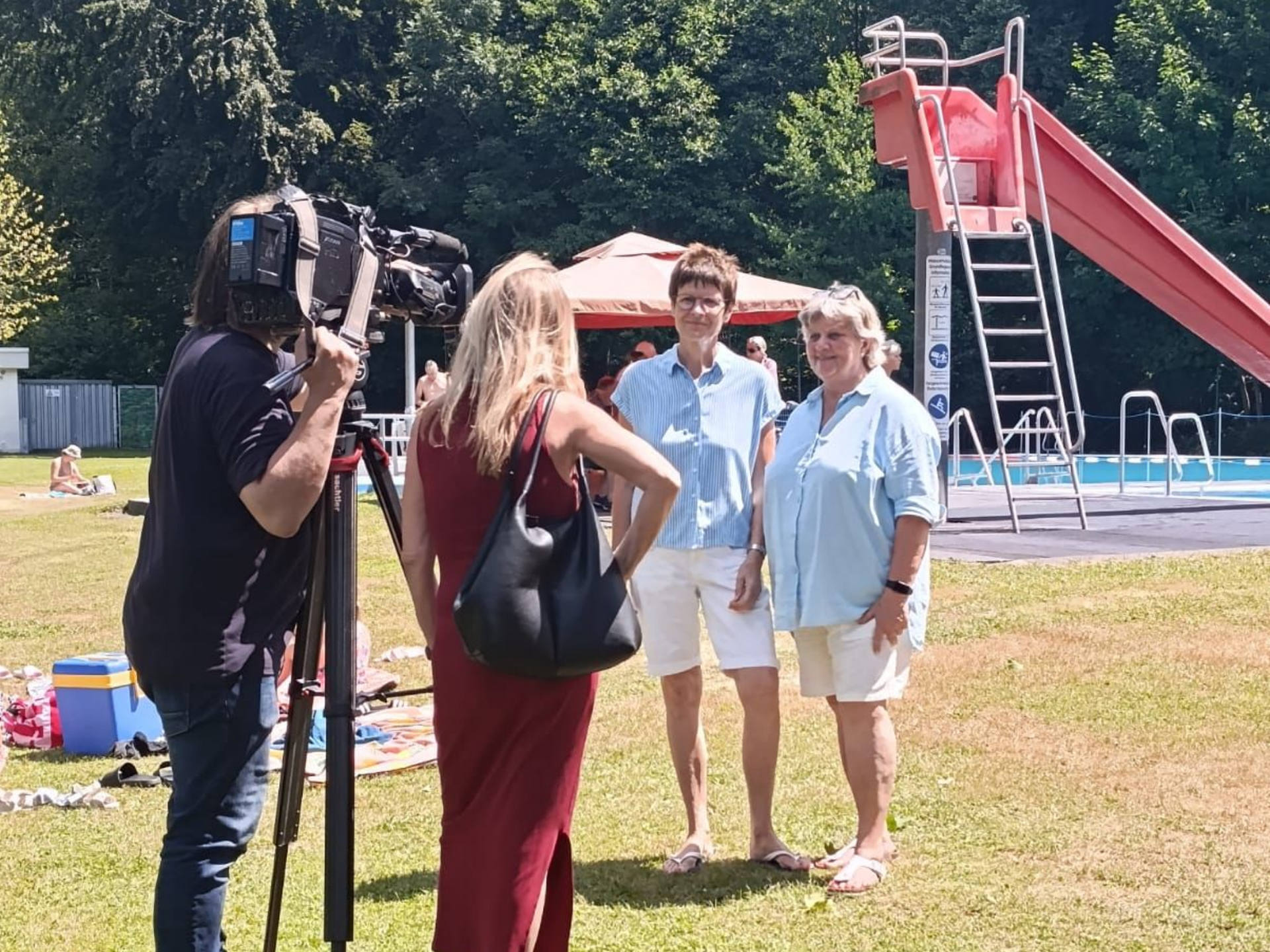 Camera crew interviewing two people near a pool with a red slide in the background