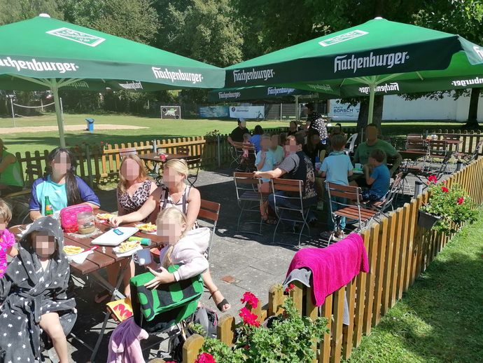 Sitzplätze im Freien mit Gästen unter grünen Sonnenschirmen neben einem Holzzaun und Blumenkästen.