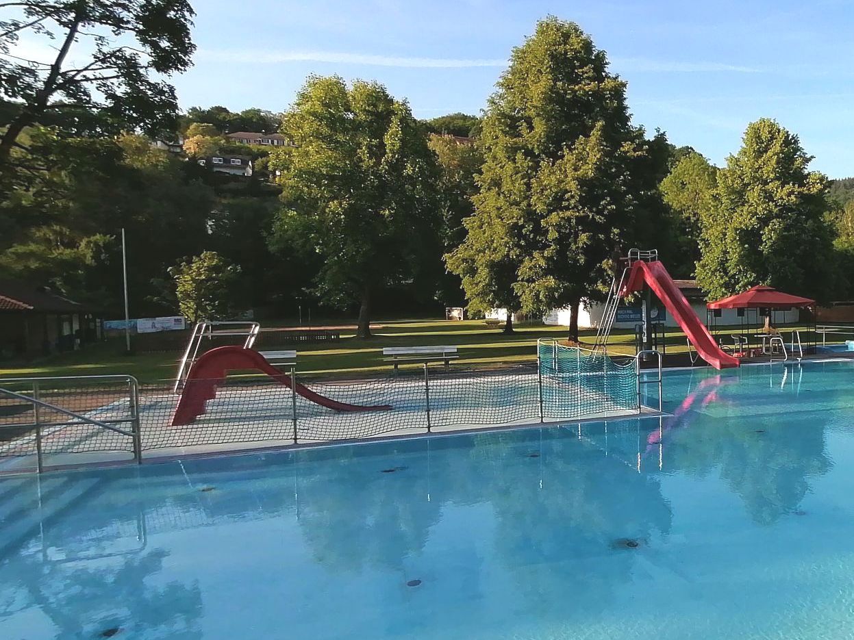 Ein Panoramabild des Freibades Oberbieber. Eine Wiese links, eine rote Rutsche in der Mitte und mehrere Schwimmbecken rechts.